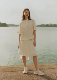 Model standing outdoors near water wearing a white polo shirt, pleated skirt, and white sneakers under warm sunlight.