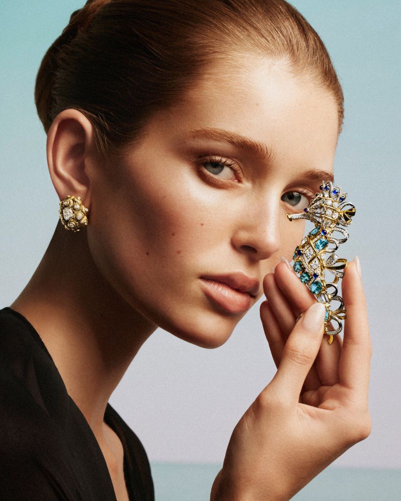 Close-up of a woman with light skin and blonde hair wearing a black top, gold earrings, holding a seahorse brooch.