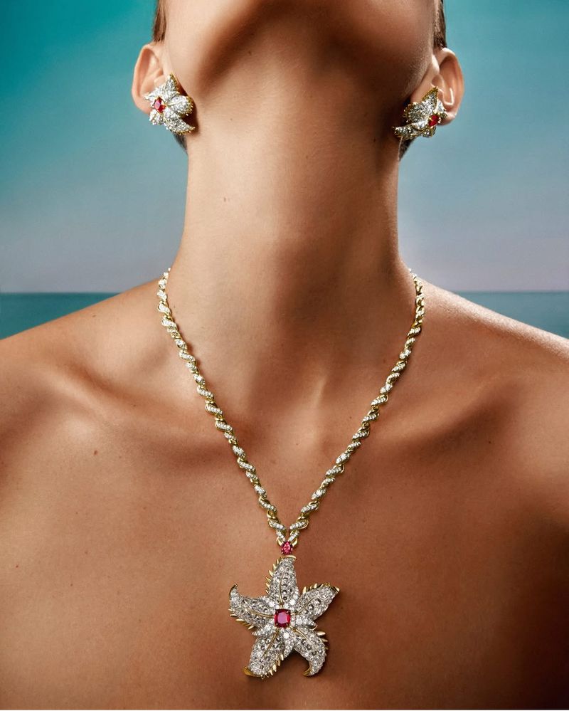 Close-up of a woman's neck and upper chest adorned with gold jewelry: a floral necklace and matching floral earrings, against an out-of-focus background.
