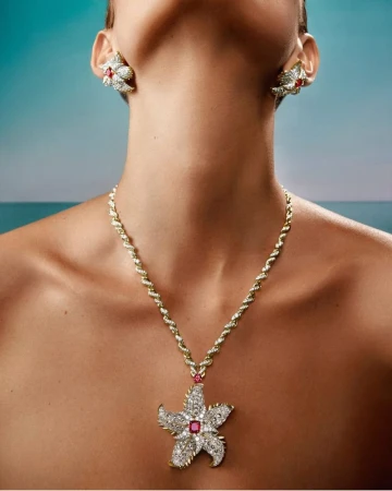 Close-up of a woman's neck and upper chest adorned with gold jewelry: a floral necklace and matching floral earrings, against an out-of-focus background.