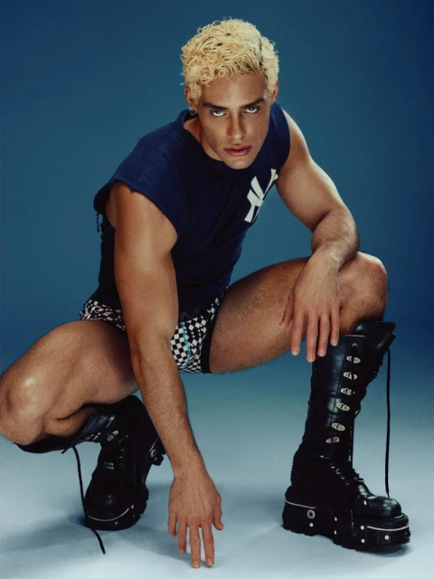 An adult male with light blonde hair crouches in heavy black boots, wearing a navy tee shirt and checkered shorts on a blue background.