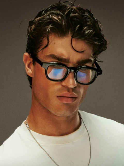 Headshot of adult male wearing black glasses and a white t-shirt with a silver chain necklace against a neutral grey background.