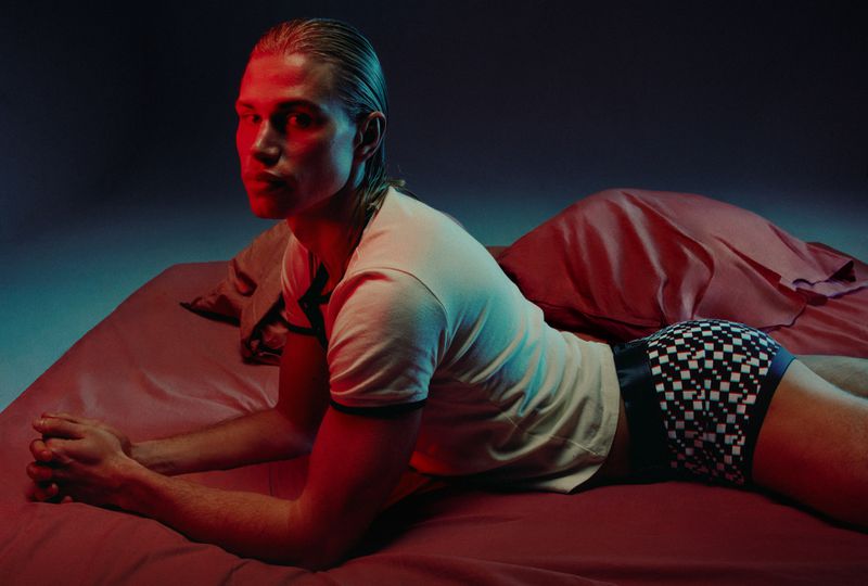 A young adult male with blonde hair poses on a bed in a photo studio with blue and red lighting, wearing a white shirt and checkered shorts.