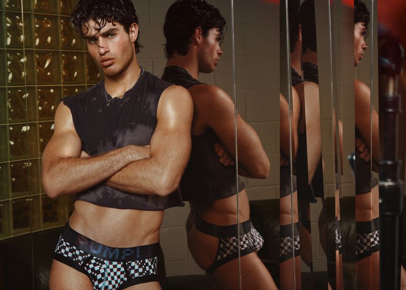 A muscular adult male poses indoors wearing a cropped black tank top and black-and-white checkered briefs in front of a mirror.
