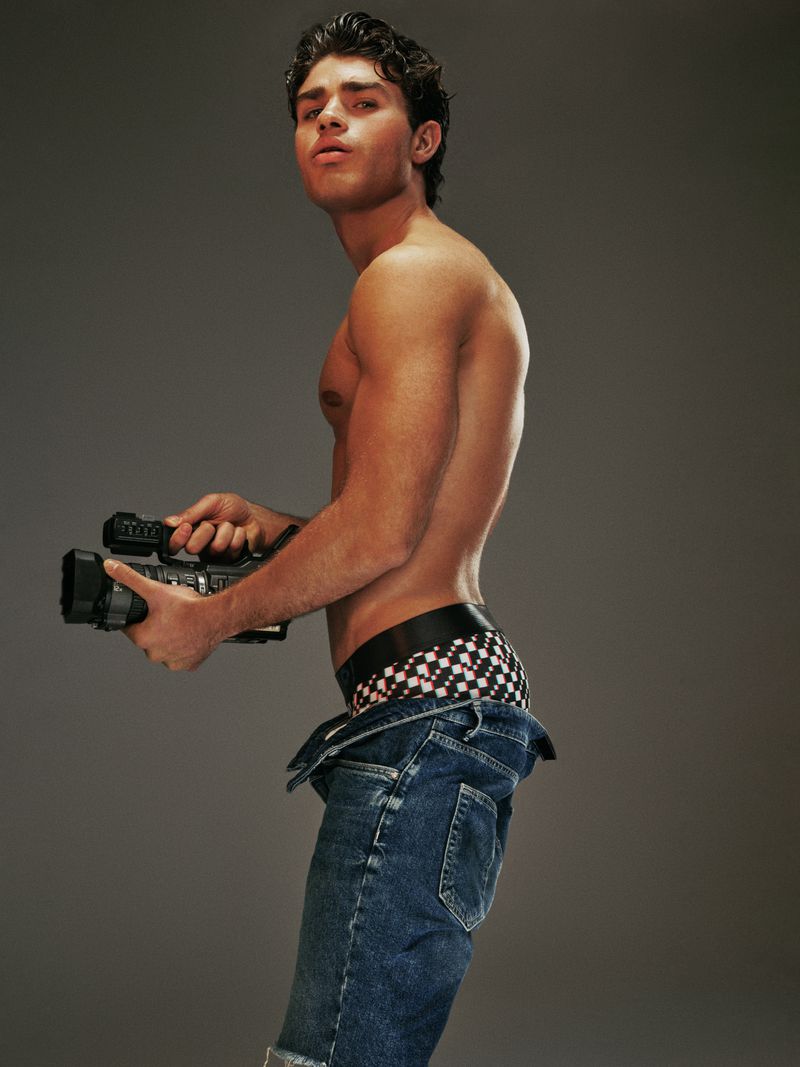 Adult male posing shirtless indoors with blue jeans, holding a vintage video camera against a solid gray background in a studio setting.