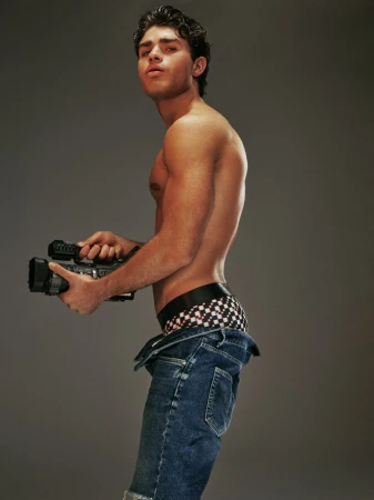 Adult male posing shirtless indoors with blue jeans, holding a vintage video camera against a solid gray background in a studio setting.
