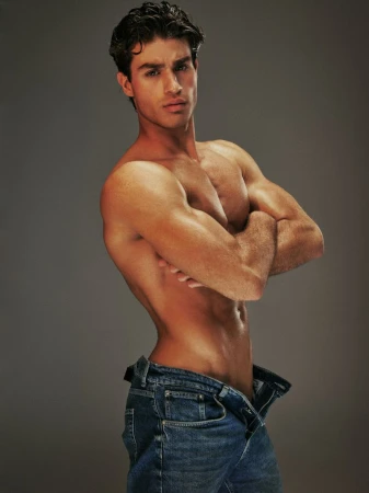Handsome male fashion model with muscular physique poses shirtless in studio with opened blue jeans and arms crossed against grey backdrop.
