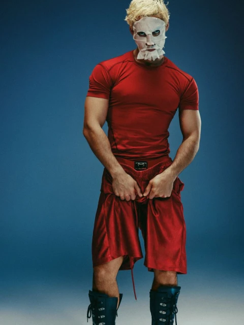 Male model wearing a red t-shirt, red shorts, and platform boots poses against a gradient blue background with a sheet mask on.