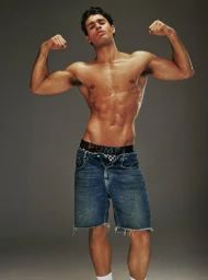 Shirtless muscular man flexing arms wearing denim shorts and branded underwear against a grey seamless studio background.