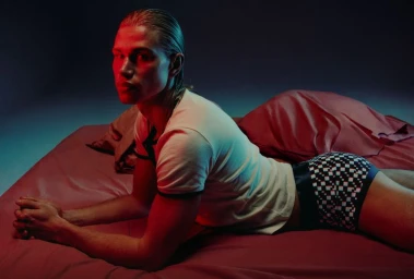 Blonde male model wearing white ringer t-shirt and patterned boxer briefs lying on a red bed under dramatic red and blue lighting.