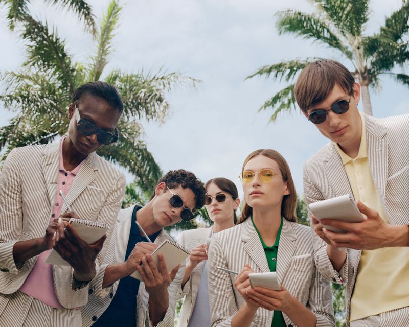 Five people wearing sunglasses and striped suits hold notebooks outdoors in a low-angle shot with palm trees against a bright sky.
