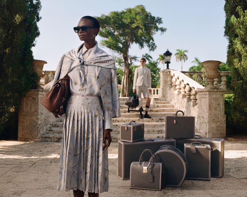 A fashionable woman in a printed suit stands outdoors with luxury luggage; a man is visible in the background near a stone building.