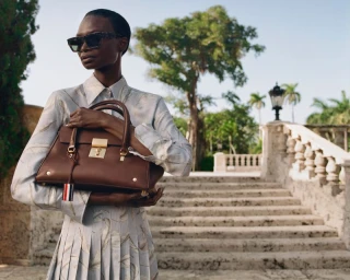 Model wearing patterned shirt and sunglasses holding a brown leather handbag outdoors near stone steps.