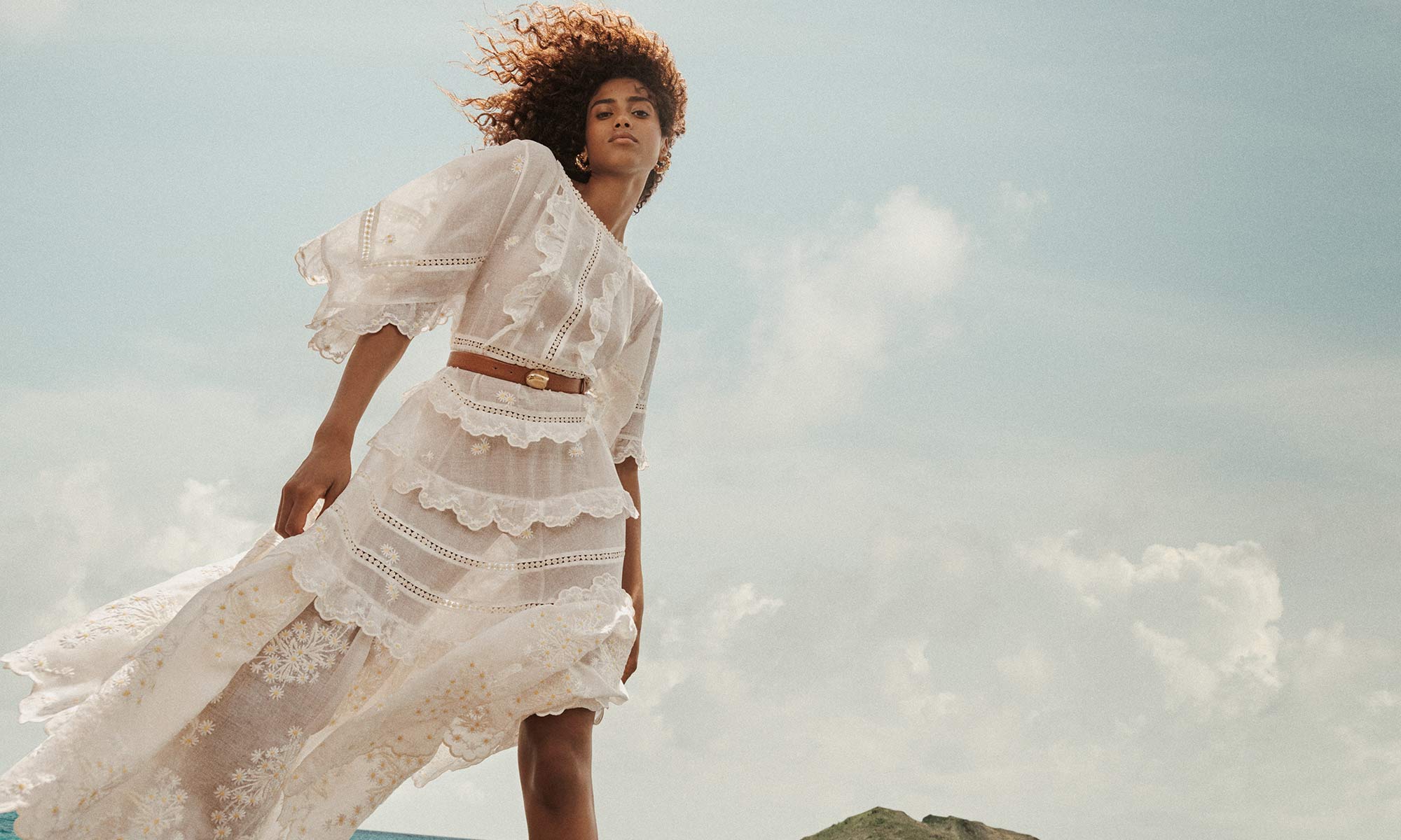 A woman in a flowing white dress poses outdoors against a sky backdrop. She wears a brown belt and her brown hair is windswept.