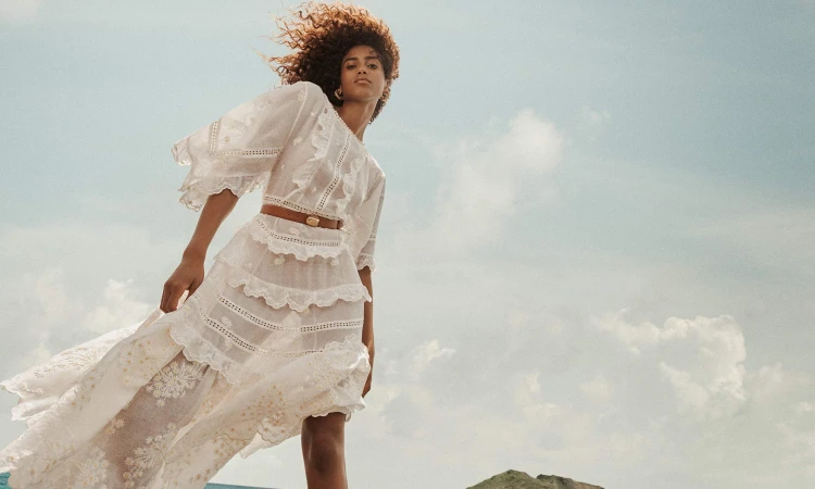A woman in a flowing white dress poses outdoors against a sky backdrop. She wears a brown belt and her brown hair is windswept.