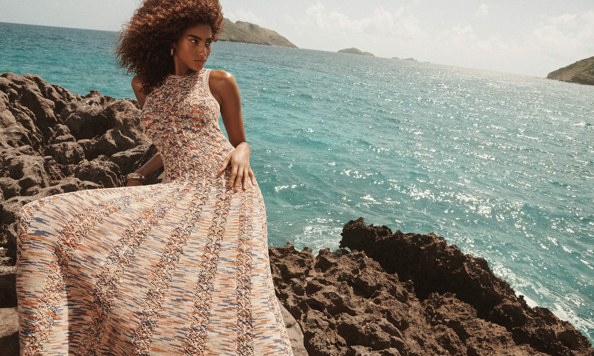 A tan-skinned woman with curly hair poses in a patterned gown on rocky terrain near a turquoise ocean with a tropical island backdrop.
