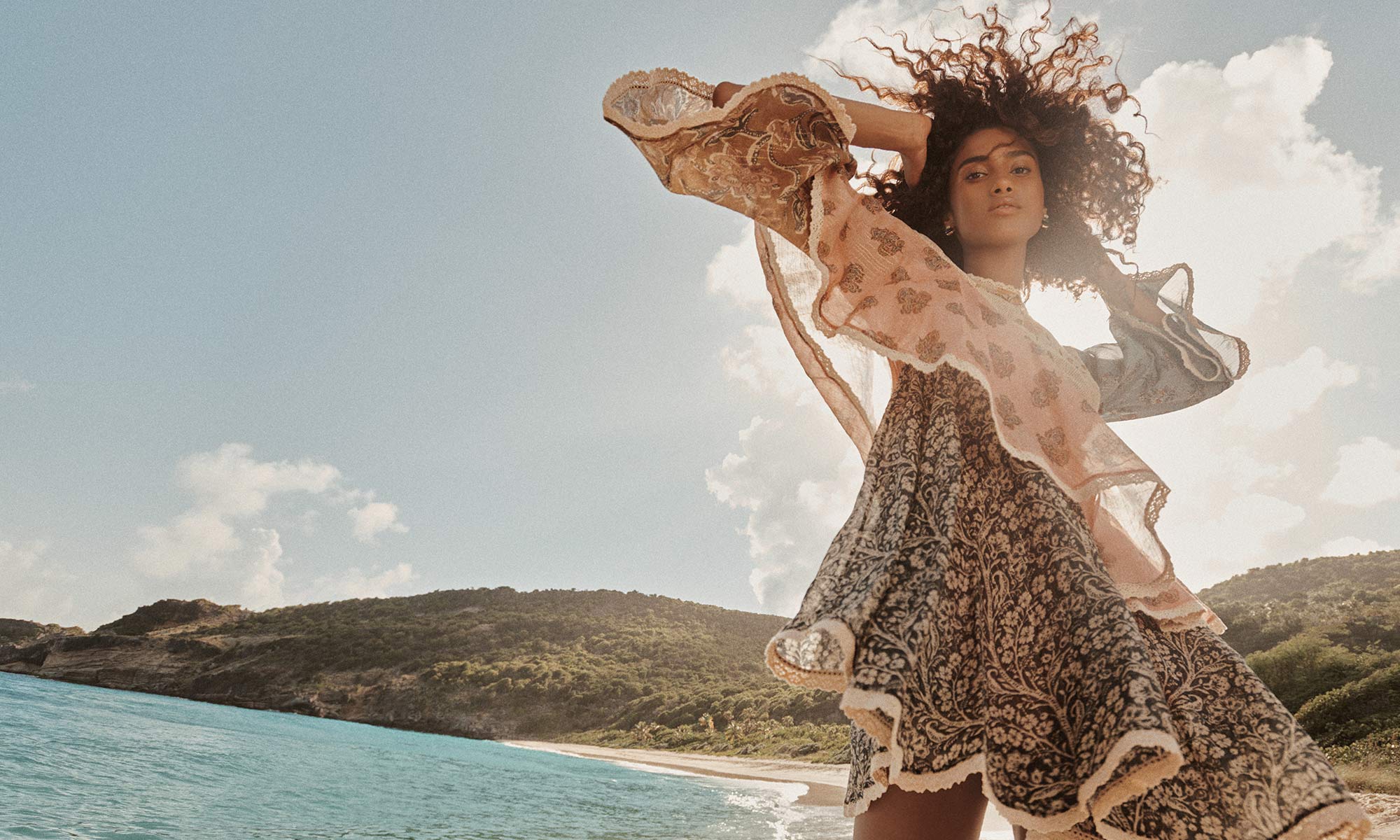 A stylish woman with curly hair poses on a beach, wearing a patterned dress with flowing sleeves against a backdrop of the sea and sky.