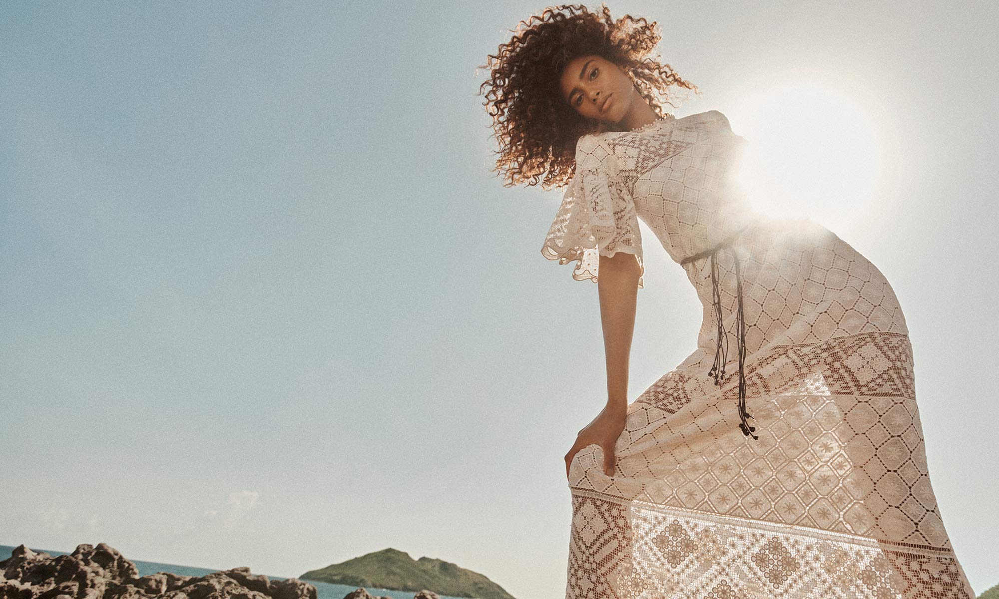 Model in a cream lace dress standing on rocky coast against a bright sky for Zimmermann Resort Swim 25