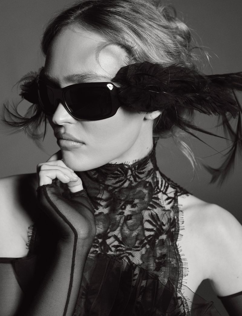 Black and white portrait of a fair-skinned adult woman wearing sunglasses with black feathers and a black lace top with long gloves.