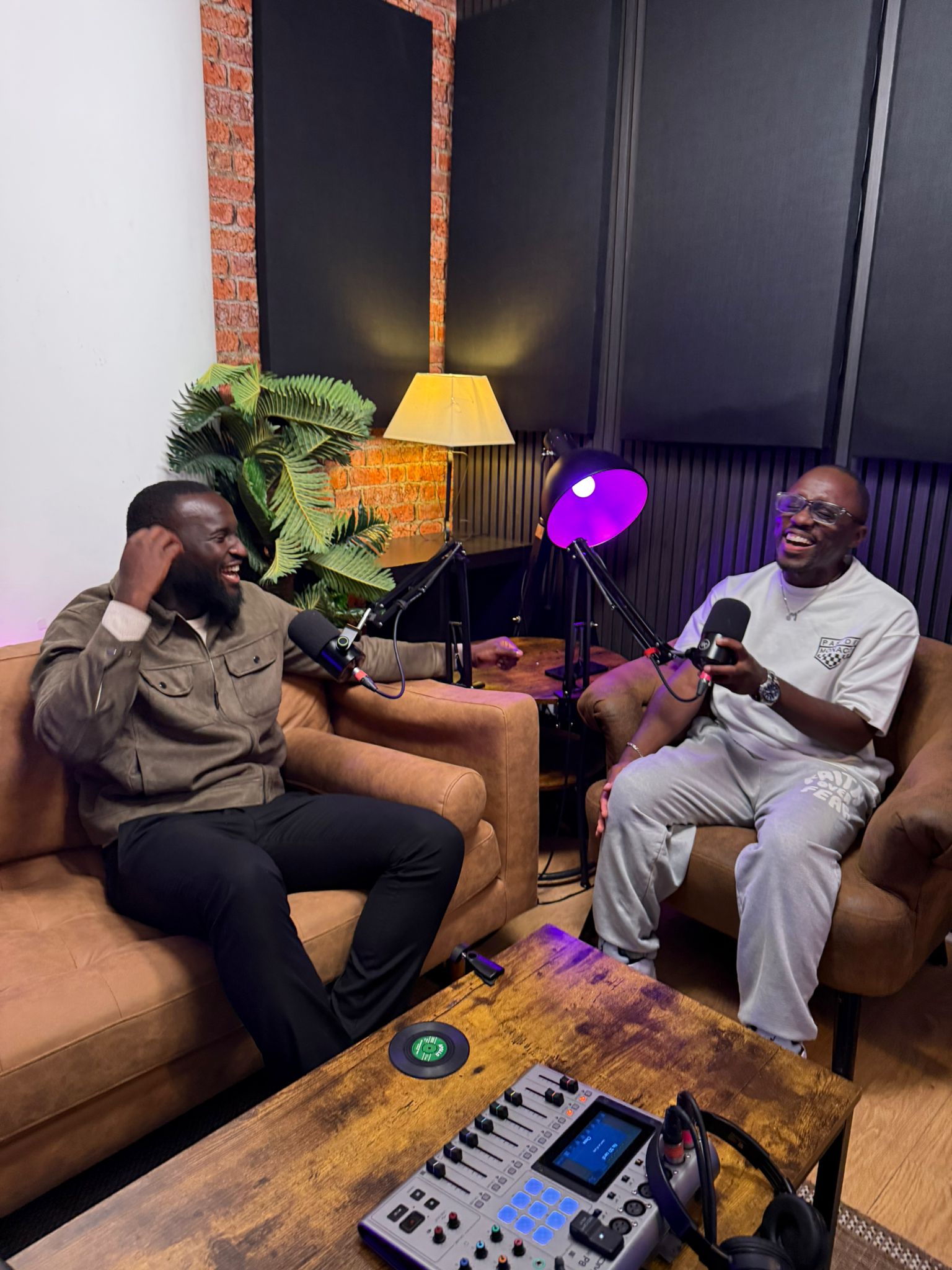 Two adult men sitting on couches in a podcasting studio, both holding microphones and talking, with sound equipment on a table.