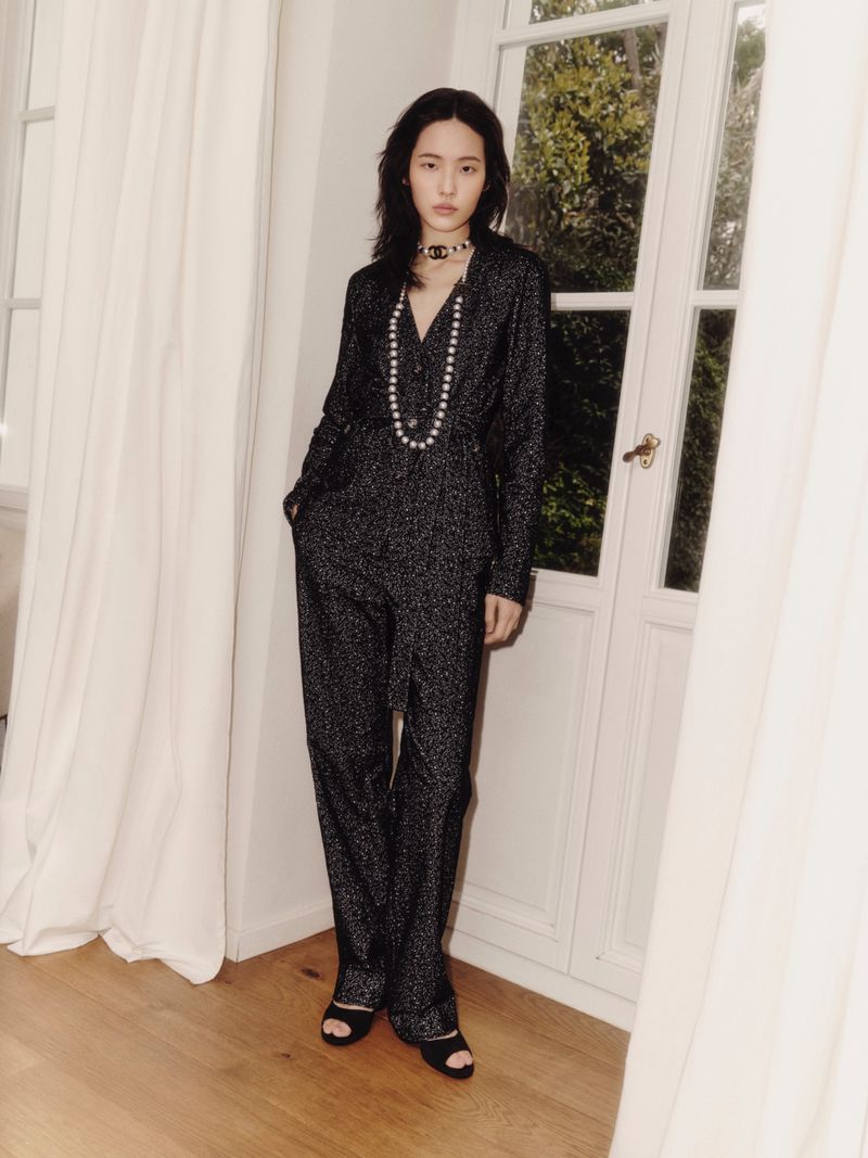 Fashion portrait of a young Asian woman in a dark, shimmery jumpsuit, layered pearl necklaces, and black heels against a white door backdrop.