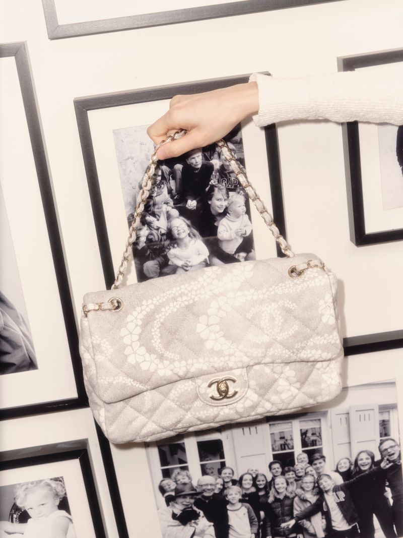 A beige Chanel handbag is suspended by an arm in front of a gallery wall displaying framed, black-and-white family photos.