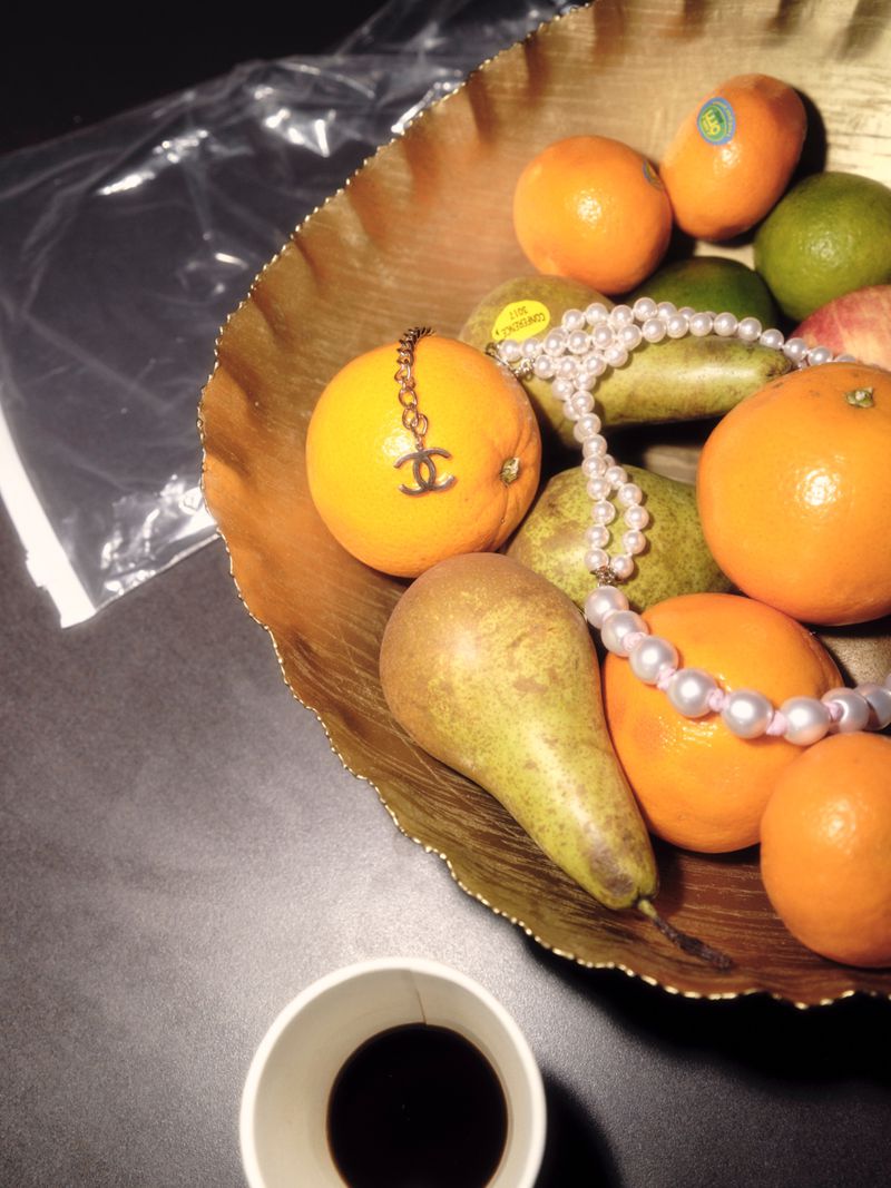 Bowl of fruit featuring oranges, pears, limes, and a strand of pearls alongside a paper cup with coffee on a black surface.