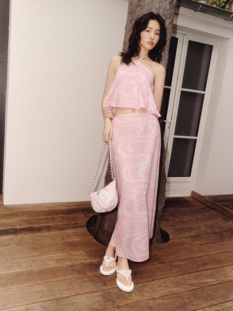 Young woman poses outdoors wearing a pink halter top and matching skirt, accessorized with a purse and sandals. Tree and building in the background.