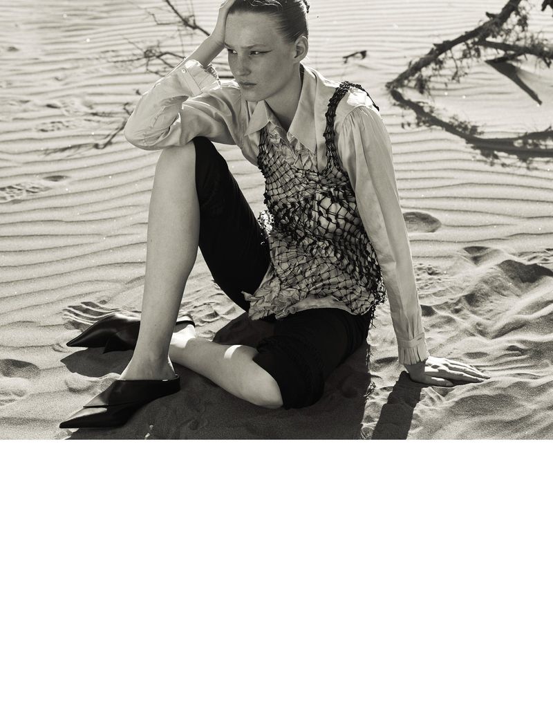 Monochrome fashion shot of a female model in a patterned top and dark shorts sitting on a sand dune with sparse branches.