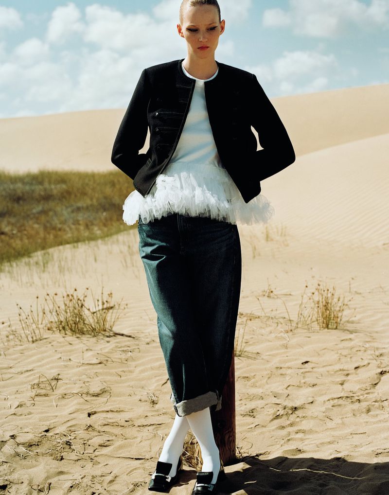 Woman in a black jacket, white top with tulle, and denim culottes stands against a desert backdrop with a wooden pole support.