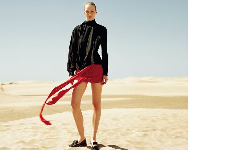 A woman in a black top and red skirt stands in a desert landscape under a clear sky, bathed in warm, natural light.