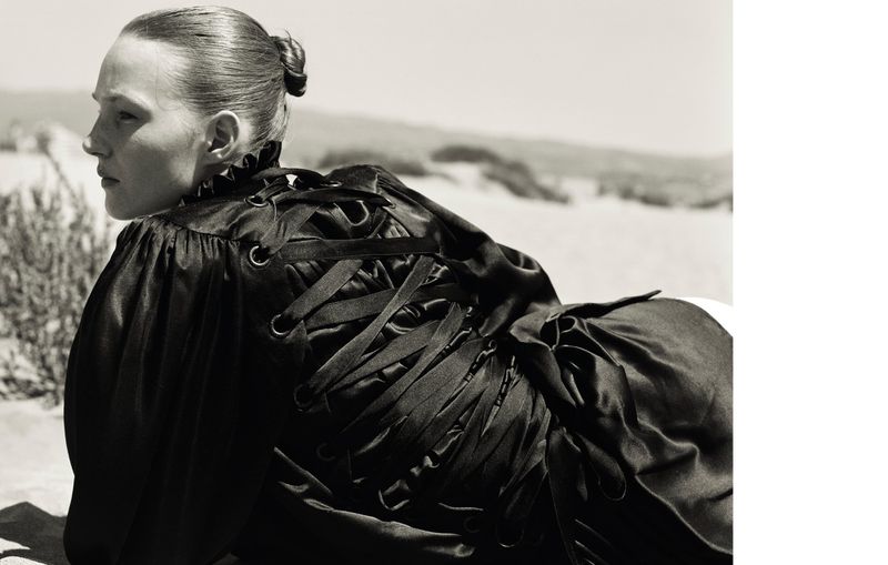 Monochrome shot of a fair-skinned woman lying down in a black dress with ribbon lacing, an outdoor setting, and high contrast lighting.