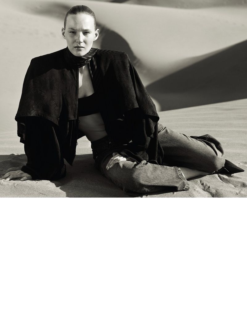 Woman in black top and jeans poses in desert dunes. Fashion editorial shot in desaturated style with neutral color palette.