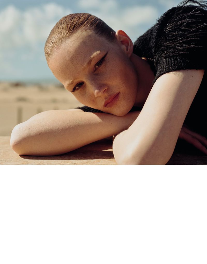 Close-up of a fair-skinned woman with dark eyeliner lying on her arms outside in a black knit top with beach and sky background.