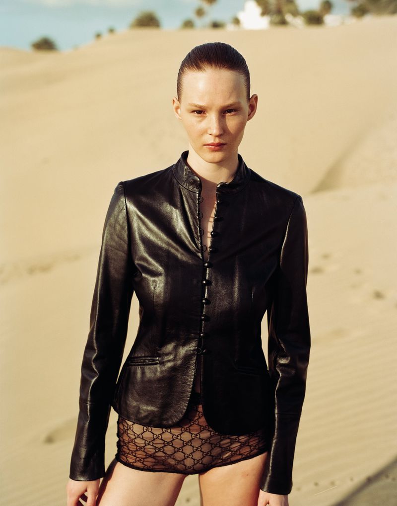 A solitary female model poses in a desert landscape, wearing a black leather jacket and patterned bottoms under the warm, natural sunlight.