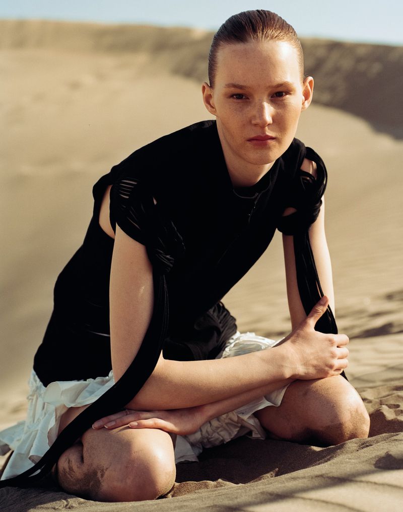 A female fashion model poses kneeling in a stark desert wearing a dark, sleeveless top with tied straps and a short, layered white skirt.