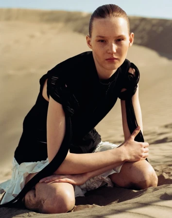 A female fashion model poses kneeling in a stark desert wearing a dark, sleeveless top with tied straps and a short, layered white skirt.
