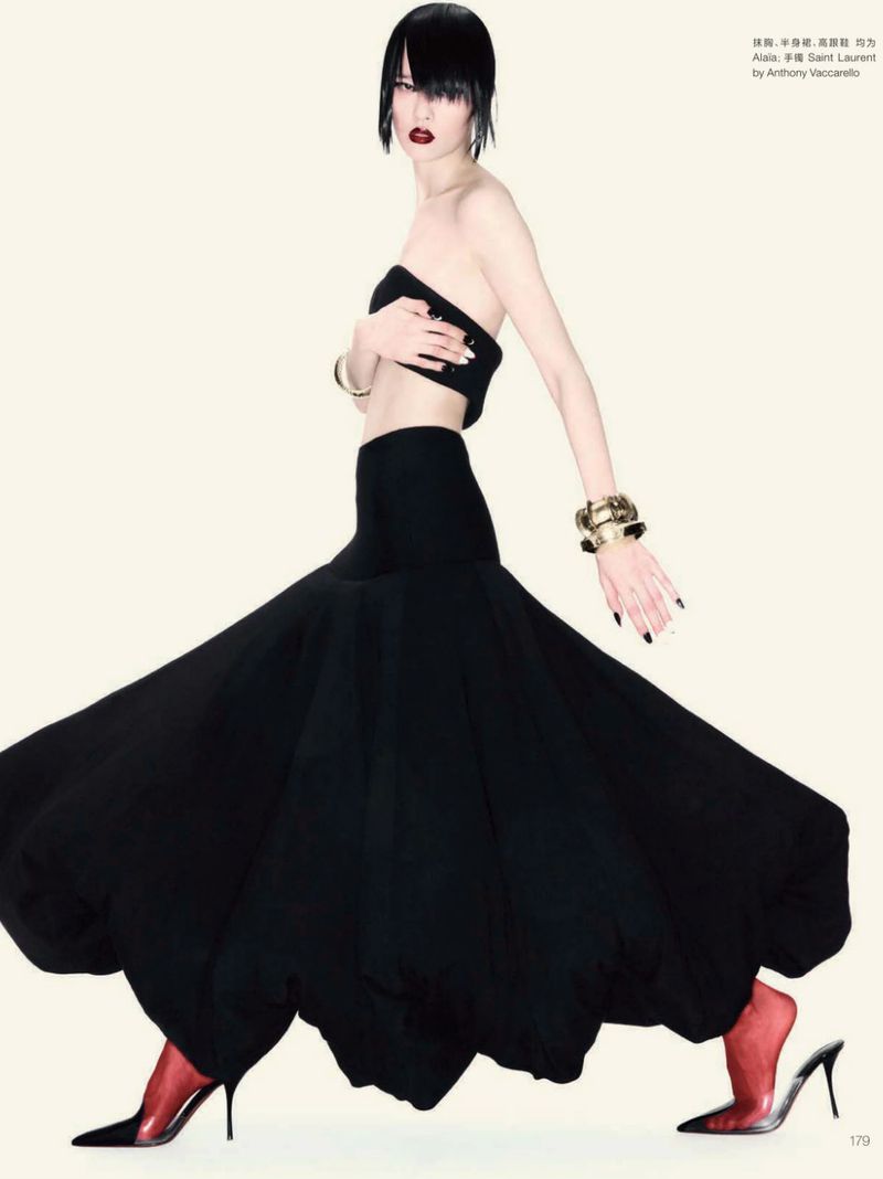 A stylish woman poses in a black tube top, long black skirt, and red heels against an off-white background, showcasing a high-fashion editorial look.