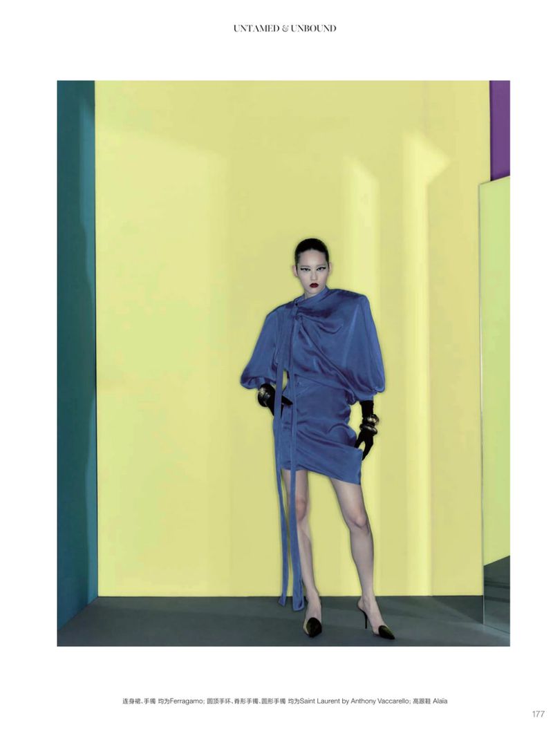 Fashion editorial shot of a woman in a blue draped mini-dress and black gloves posing against a yellow and green wall with a purple accent.