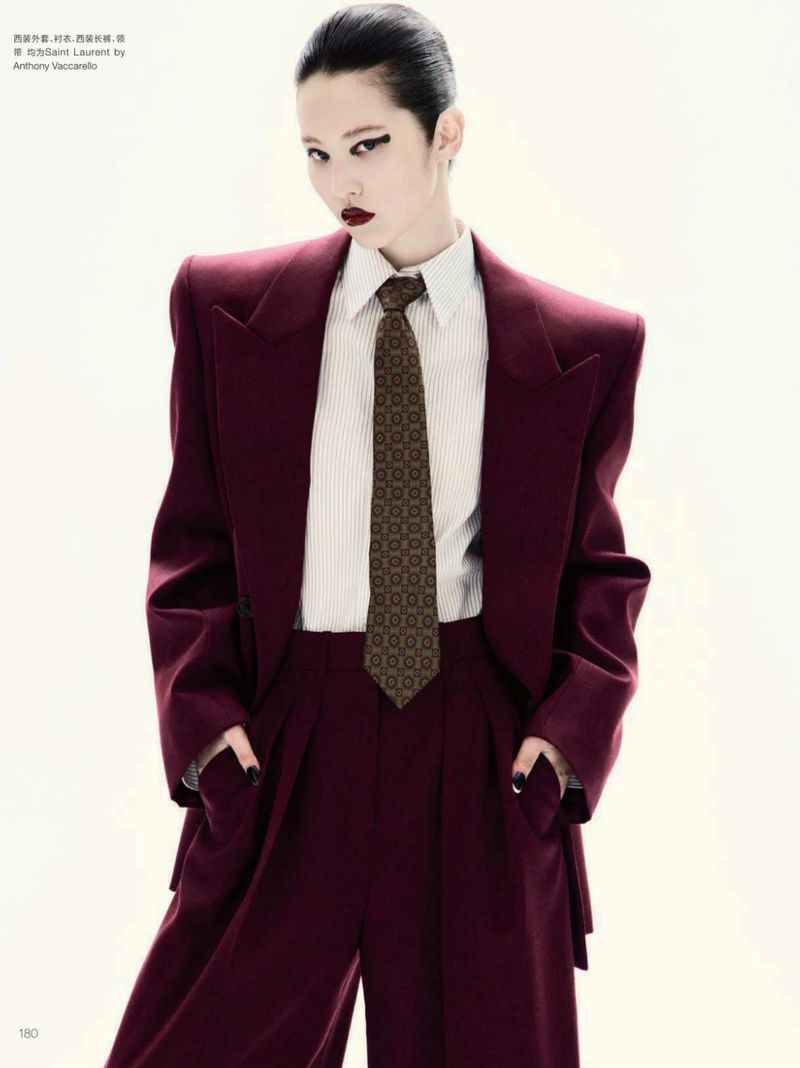 Adult female model in a maroon suit and tie posing in a studio with a neutral background for a fashion editorial style photograph.