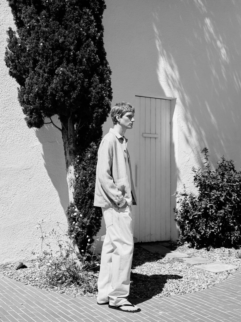 Monochrome full shot of a blonde adult man wearing light-colored casual jacket and pants, standing in front of a white wall near a tree.