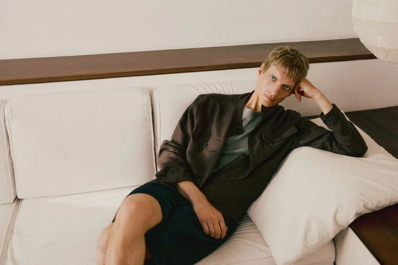 Adult male lounges on a white couch in casual wear, featuring a brown button-up shirt and dark shorts, with soft indoor lighting.