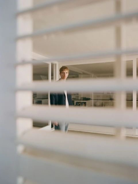 Blond man in blue jacket stands indoors, partially obscured by white window blinds in a bright room with natural light.