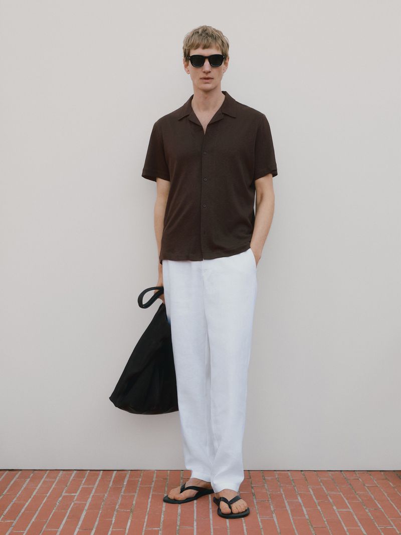 A man in a brown shirt and white pants stands on a brick surface with a black bag, wearing sunglasses and sandals for a fashion shot.