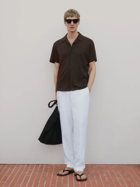 A man in a brown shirt and white pants stands on a brick surface with a black bag, wearing sunglasses and sandals for a fashion shot.