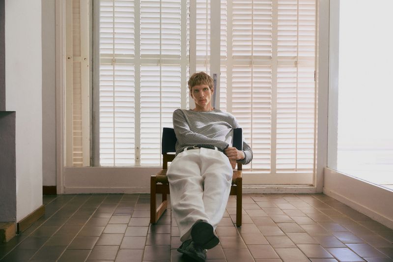 Blonde man sits in a chair indoors near a bright window. He wears a striped long-sleeve shirt and white pants. Neutral color palette.