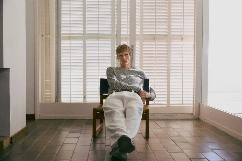 Blonde man sits in a chair indoors near a bright window. He wears a striped long-sleeve shirt and white pants. Neutral color palette.