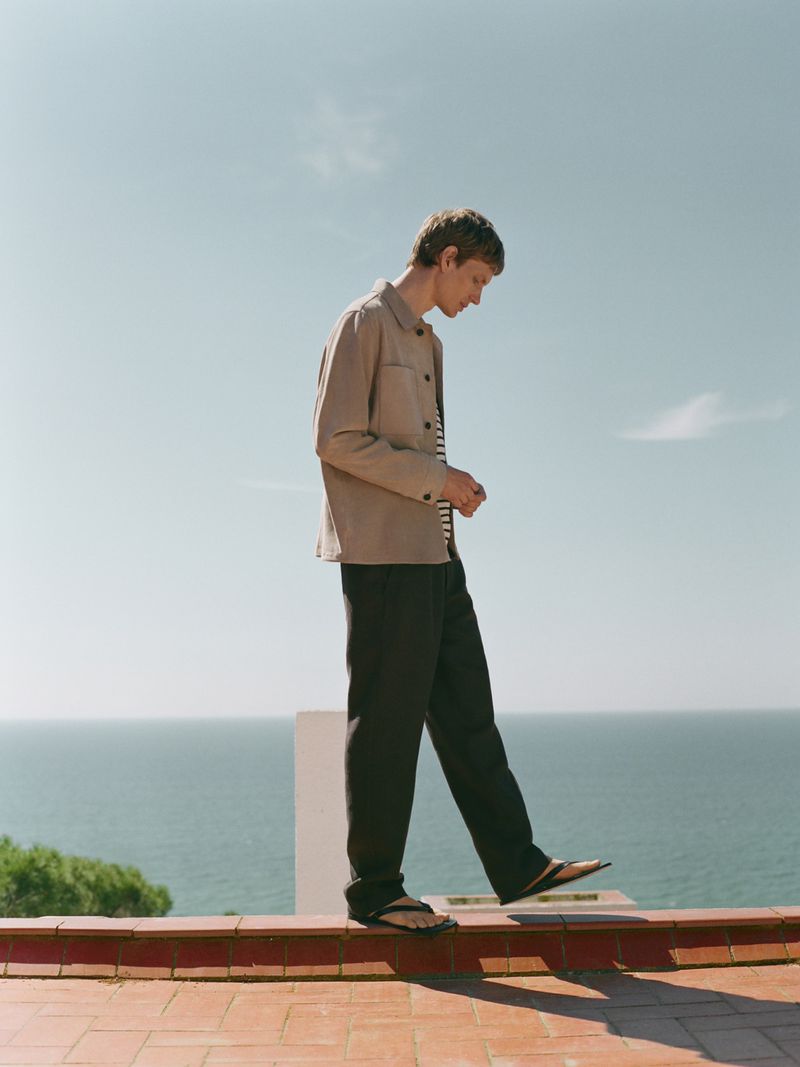 A young adult male in beige jacket and dark pants balances on a low brick wall against a background of blue sky and the ocean.