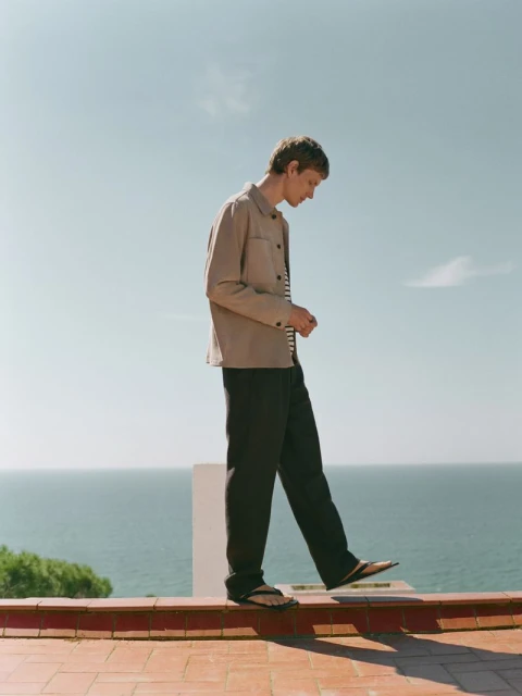 A young adult male in beige jacket and dark pants balances on a low brick wall against a background of blue sky and the ocean.