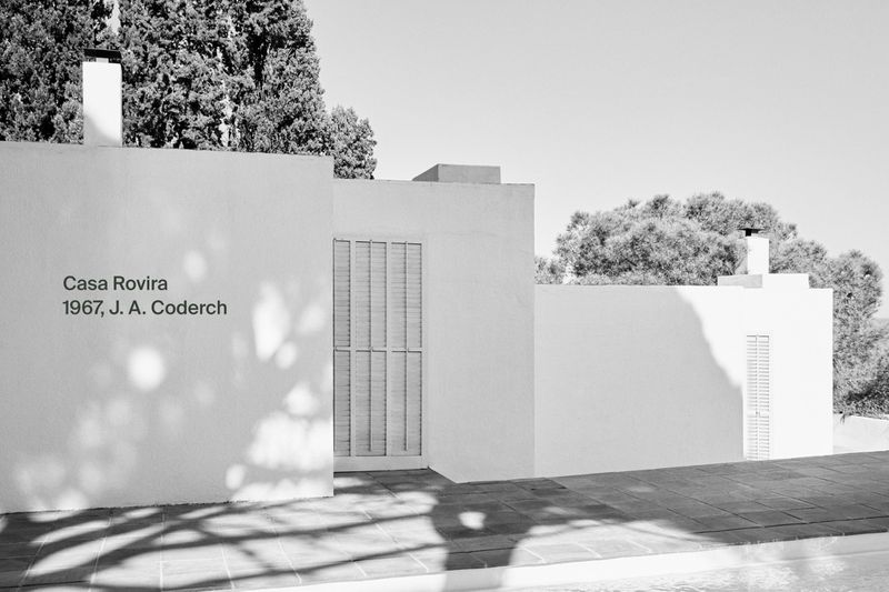 Architectural image of the Casa Rovira building with a white facade, closed shutters, and a flat roof, partially shaded by nearby trees.